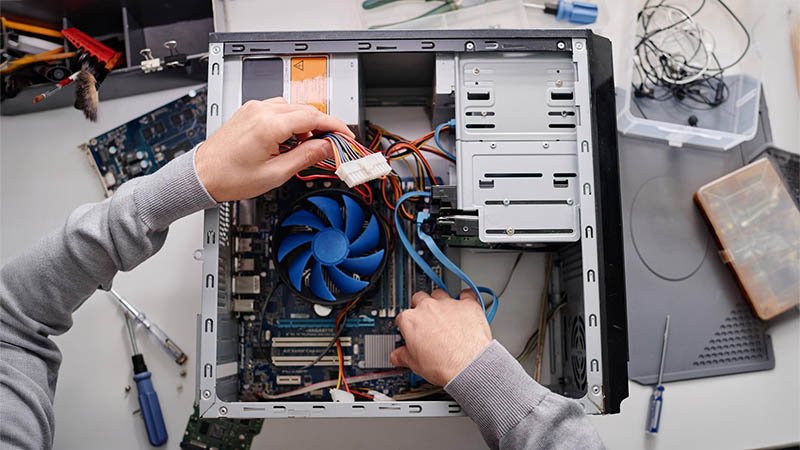 COMPUTER WEB A person is working on the internal components of an open desktop computer. Visible hardware includes the motherboard, a large blue CPU fan, power supply unit, and multiple cables. One hand is connecting a power cable, while another adjusts a component on the motherboard. Surrounding the computer are tools and electronic parts such as screwdrivers, wires, and circuit boards, indicating a hands-on repair or custom PC assembly process.