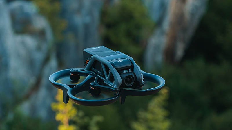 DRONE WEB A DJI Avata drone hovers mid-flight outdoors against a blurred backdrop of trees and rocky cliffs. The compact quadcopter features a front-mounted camera and protective guards around its propellers, designed for immersive FPV (First Person View) flying. Its rugged body and stabilized design suggest suitability for aerial photography, videography, and recreational use in natural environments.