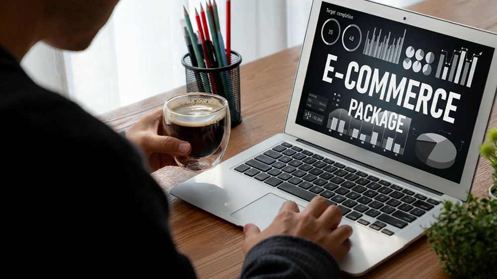 Person sitting at a wooden desk, holding a glass cup of coffee and using a laptop displaying charts and graphs labeled “E-COMMERCE PACKAGE,” with progress indicators showing 35% and 48%. A black mesh pencil holder with colored pencils sits nearby.